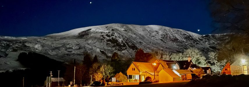 Glen Clova Hotel at night with snowcapped mountains and a starry night behind the hotel lit up in the darkness