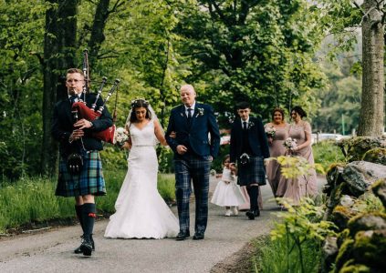 A bride with her father and bridesmaids following a lone piper down a country lane to her wedding at Glen Clova