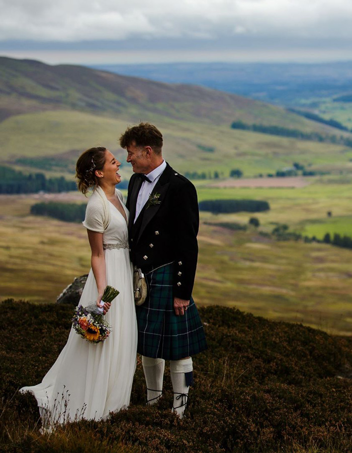 A married couple laughing on a Heather moorland in Glen Clova with rolling hills and a valley behind them. Photography by Barry Robb Photography. https://barryrobbphotography.com/